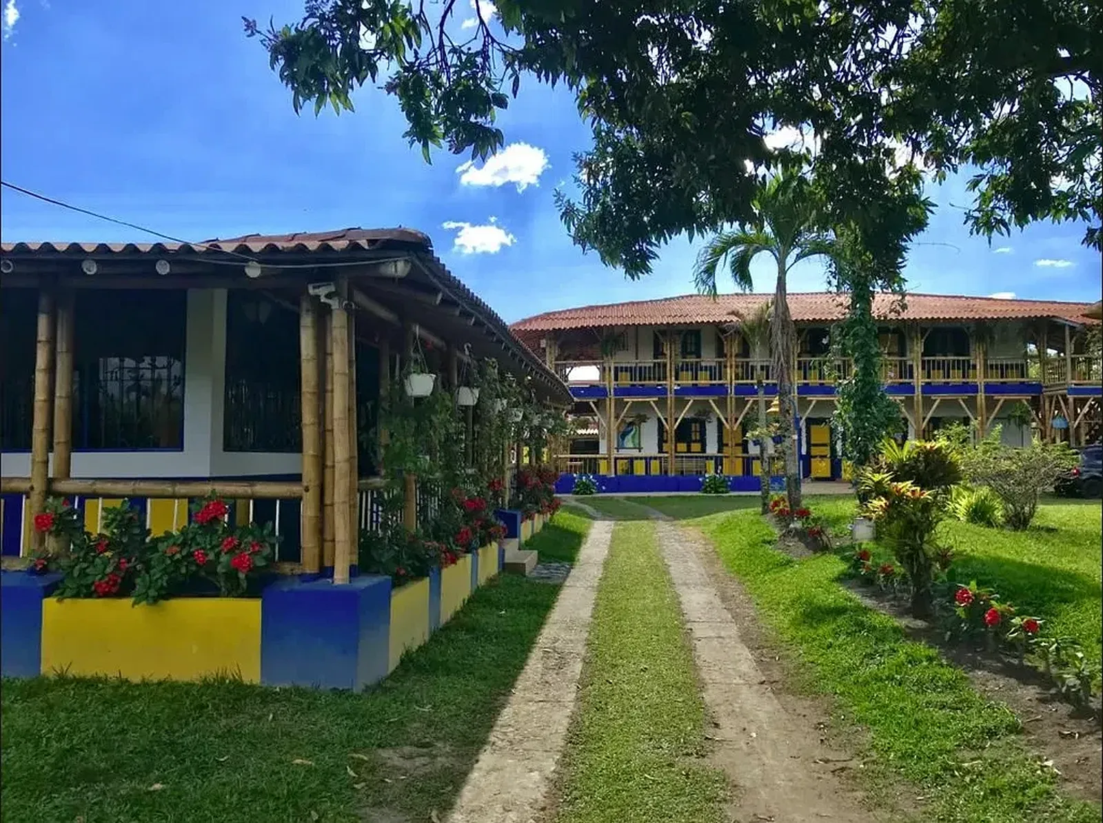 Casona de guadua de dos pisos, pintada de azul y amarillo, con camino de piedra entre el jardín